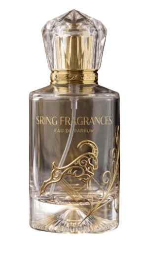 A clear glass perfume bottle with a faceted clear cap and ornate gold lettering reading 'SRING FRAGRANCES' and 'EAU DE PARFUM'. The bottle has a gold goat motif printed on the front and is photographed against a warm blurred brown background on a reflective surface.